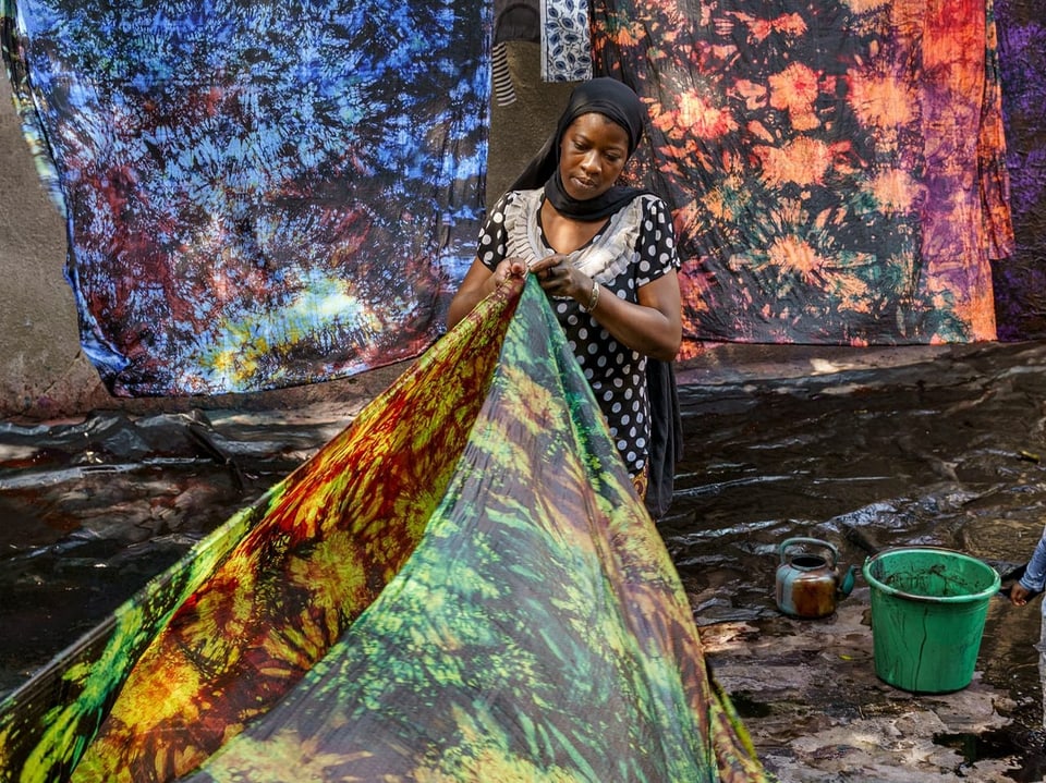 Eine Frau mit Schleier steht vor einer Wäscheleine mit bunt gefärbten Stoffen. Eine der Stoffe hält sie in der Hand.