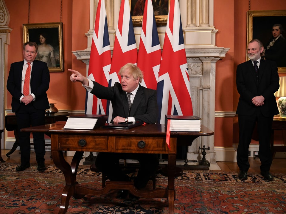 Johnson unterzeichnet Brexit-Papiere und zeigt mit der Hand triumphierend in die Ferne.