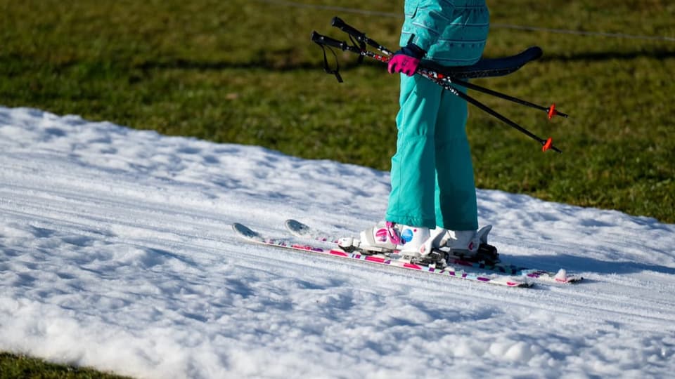 Ein Mädchen fährt auf einem Bügellift auf ihren Skiern hoch. Darunter ist eine dünne Schneeschicht, daneben grün.