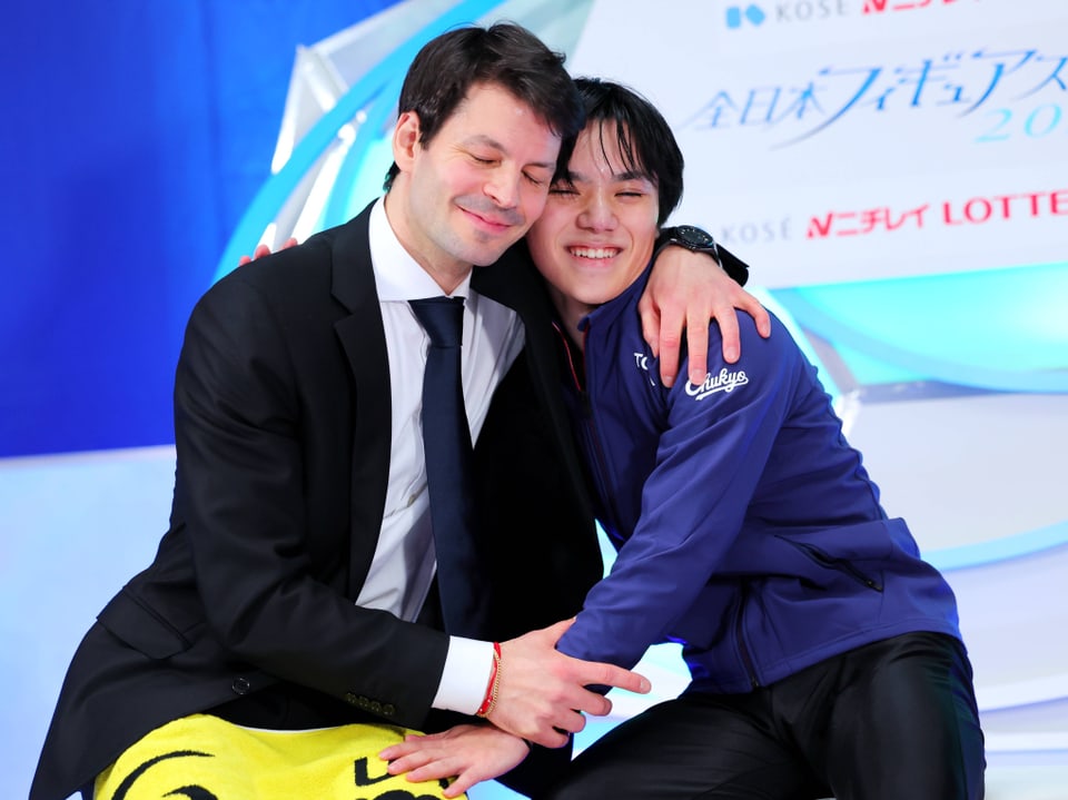 Zwei Männer in Anzügen umarmen sich lächelnd bei einer Sportveranstaltung.