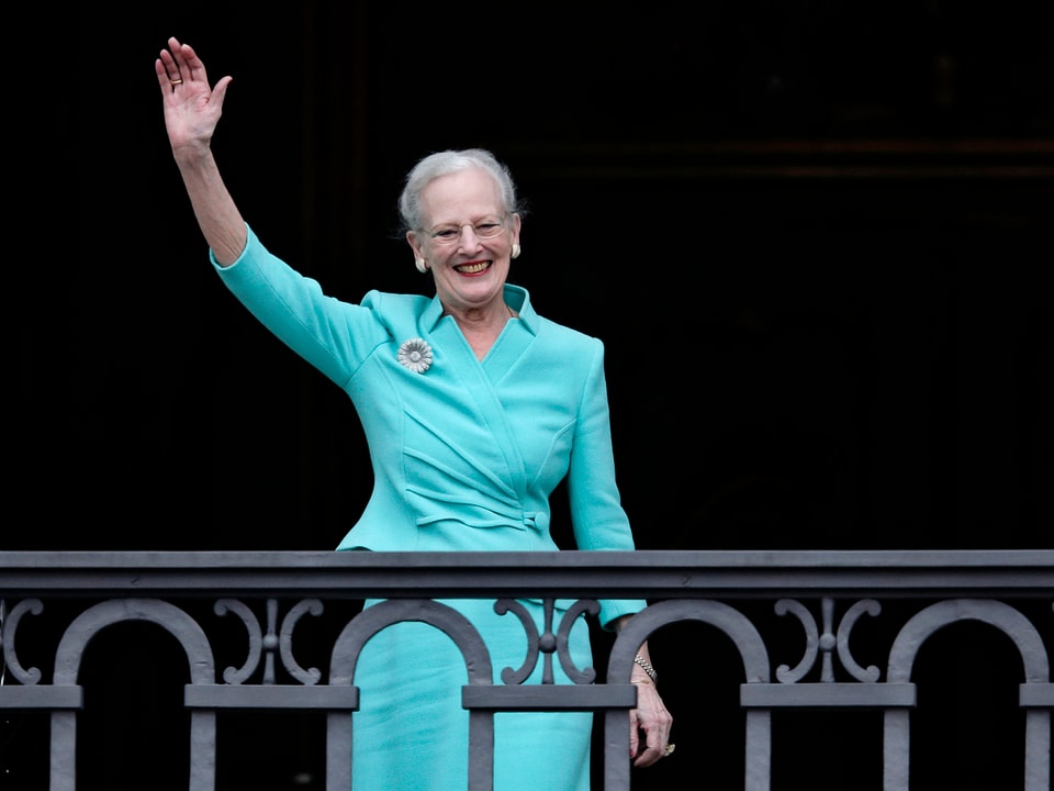 Königin Margrethe steht auf einem Balkon in einem Türkis farbenen Kleid. Sie winkt und lacht.