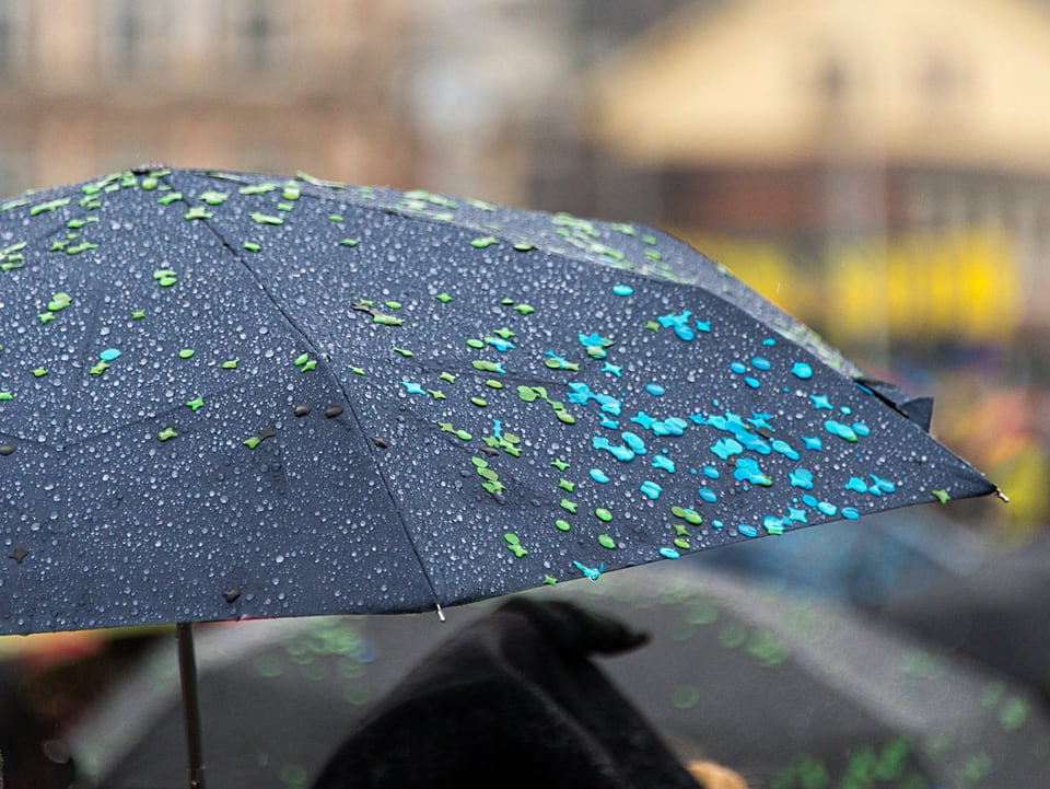 Schwarzer Regenschirm, der aufgespannt ist. Darauf kleben kleine, farbige Papierstückchen in grün und blau. 