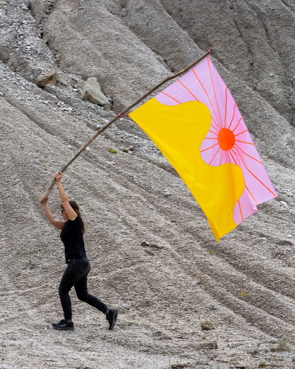 Eine Frau hält eine gelb-pinke Fahne und läuft vor einem Kies-Hintergrund. 