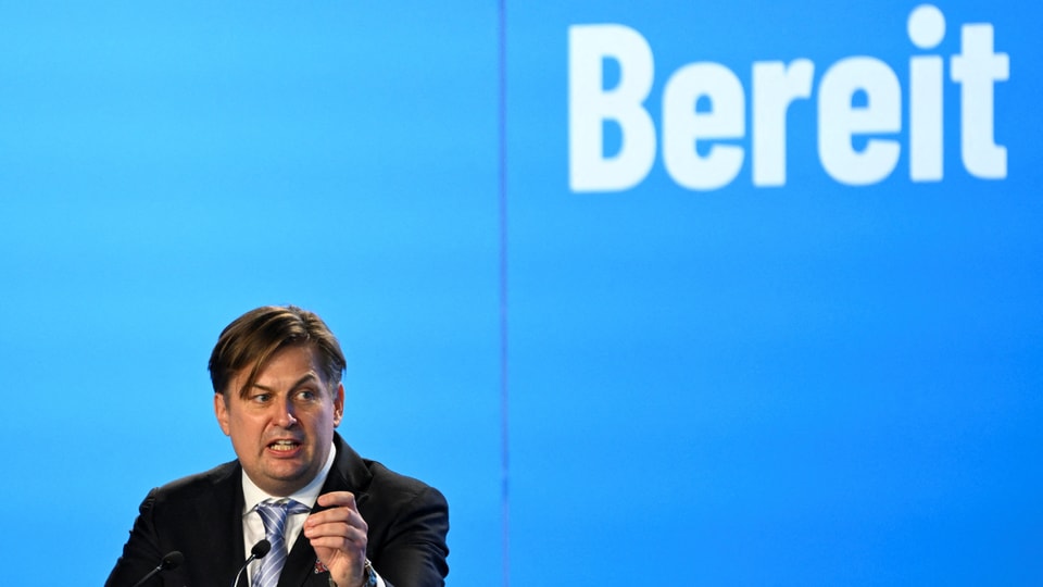 Mann in Anzug gestikuliert beim Reden vor einem blauen Hintergrund mit dem Wort 'Bereit'.