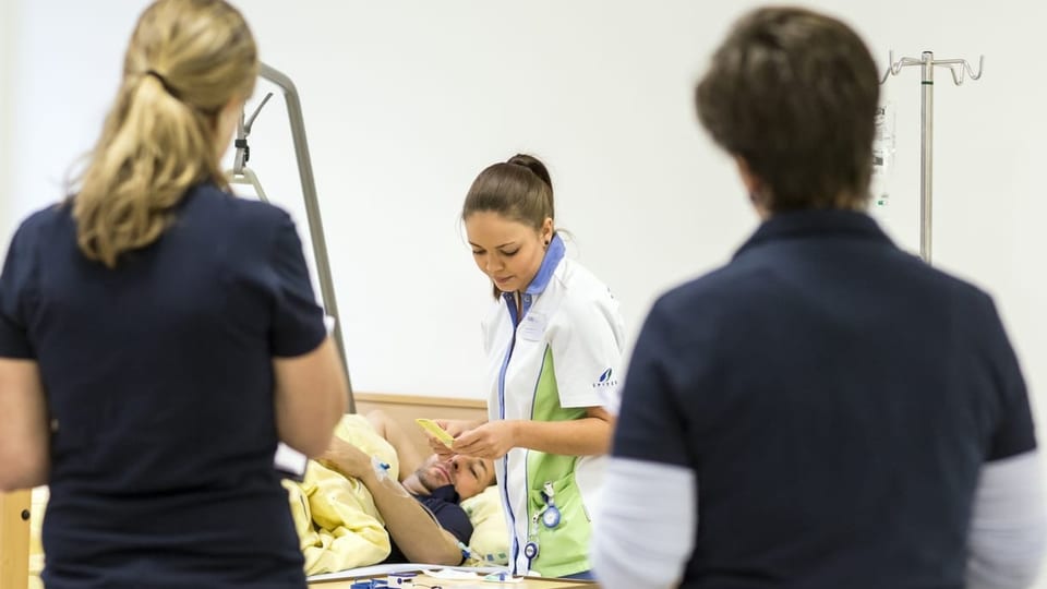 Eine Spitex-Mitarbeitende behandelt einen Patienten, zwei Frauen stehen daneben.