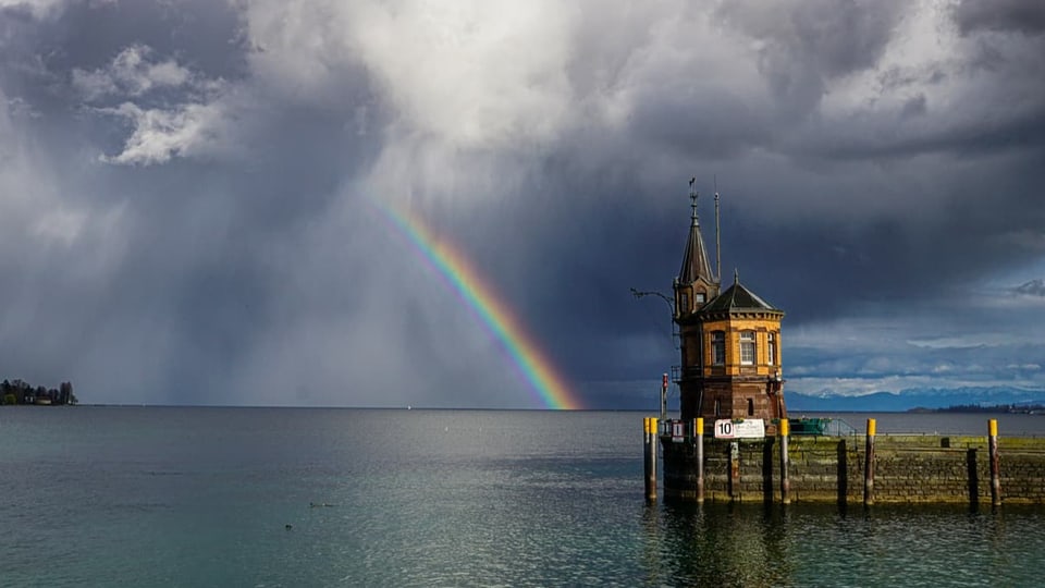 Blick vom Hafen auf einen grossen See mit dunklen Wolken und Regenbogen über dem Wasser. 