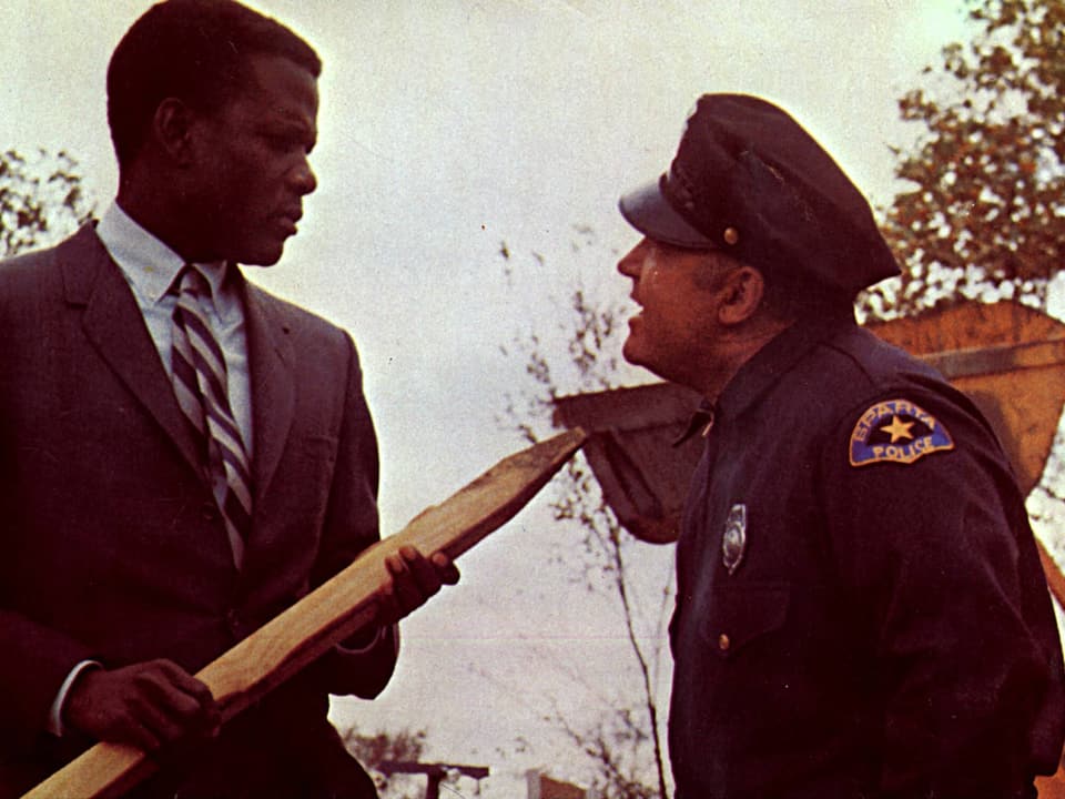 Ein dunkelhäutiger Mann mit Pfahl in der Hand neben einem Polizisten in einer Filmszene.