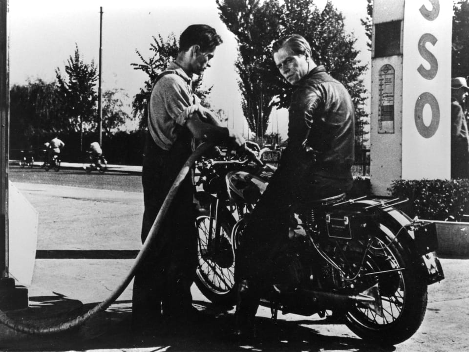 Ein Tankwart betrankt das Motorrad eines anderen Mannes, welcher auf dem Motorrad sitzt.