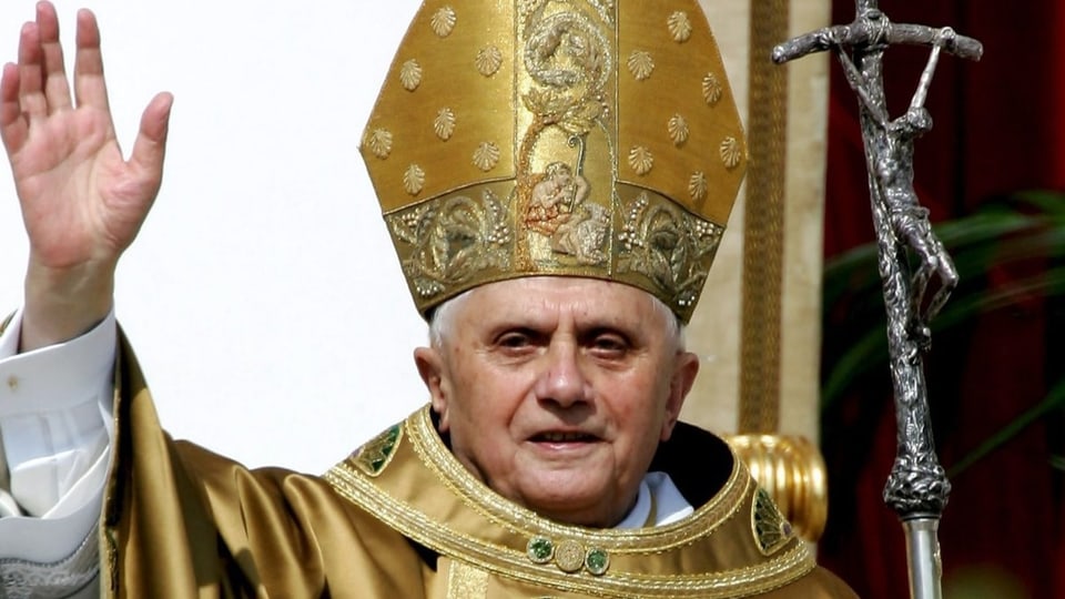 Papst Benedikt XVI. winkt und hält ein Kreuz mit Jesus in der Hand. Er trägt goldene und weisse Kleidung.