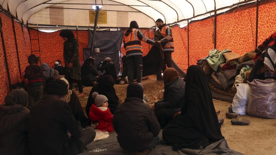 Menschen sitzen in einer Notunterkunft, hinter ihnen stehen zwei Helfer.