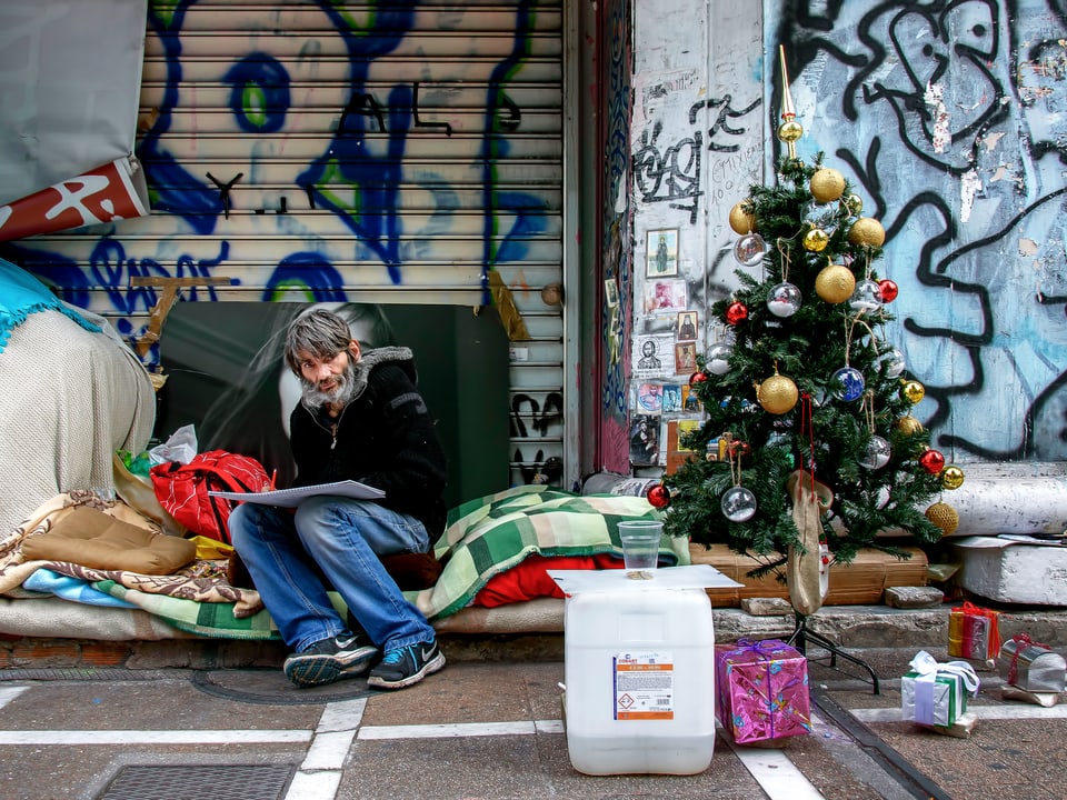 Ein Obdachloser sitzt auf einem Stapel aus Wolldecken, neben ihm ein Tannenbaum. 