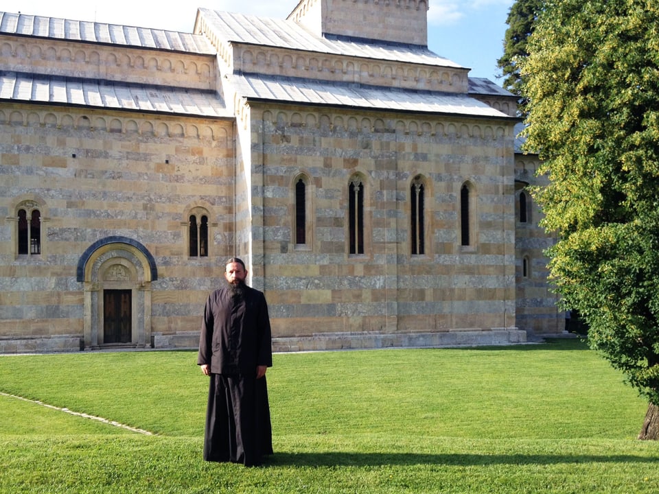 Mönch mit langem Bart und schwarzem Gewand steht im Gras vor einer Kirche.