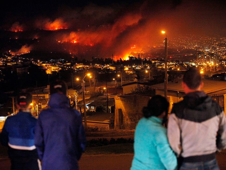 Vier Personen sehen aus der Ferne zu, wie das Feuer sich in der Stadt ausbreitet.