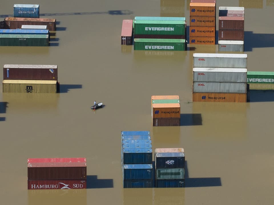 Containerhafen steht komplett unter Wasser. Mann fährt mit Boot zwischen den Containern umher.