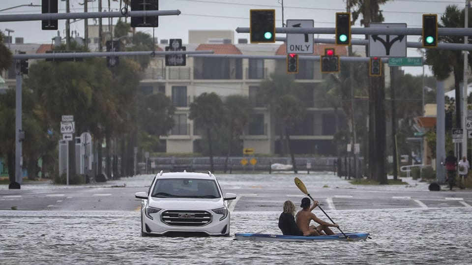 Ein Auto fährt auf einer überfluteten Strasse in Richtung Kamera. Daneben rudert ein Paar auf einem Kajak .