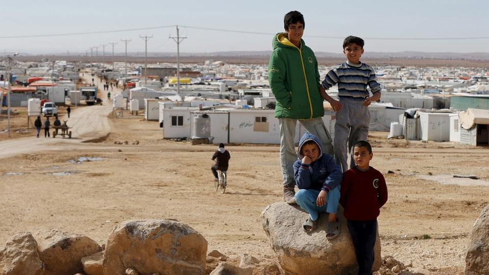 Im Vordergrund vier Kinder, im Hintergrund in wüstenähnlichem Gebiet ein Flüchtlingscamp aus Containern.