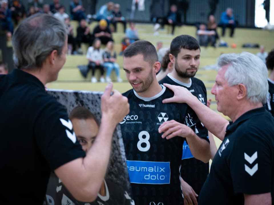 Handballspieler in Diskussion mit Trainern am Spielfeldrand in einer Sporthalle.