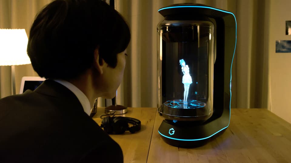 Ein junger Mann im Anzug sitzt am Tisch uns spricht zu einem Hologramm hinter der Glasscheibe.