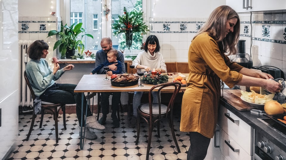 Hinten Esstisch mit drei Erwachsenen, einem Kind. vorne steht eine Frau und schneidet etwas auf Küchenbrett