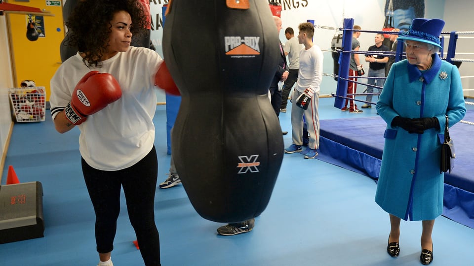 Queen Elizabeth steht in der rechten Bildhälfte. Auf der linken Seite trainiert eine Boxerin an einem Boxsack.