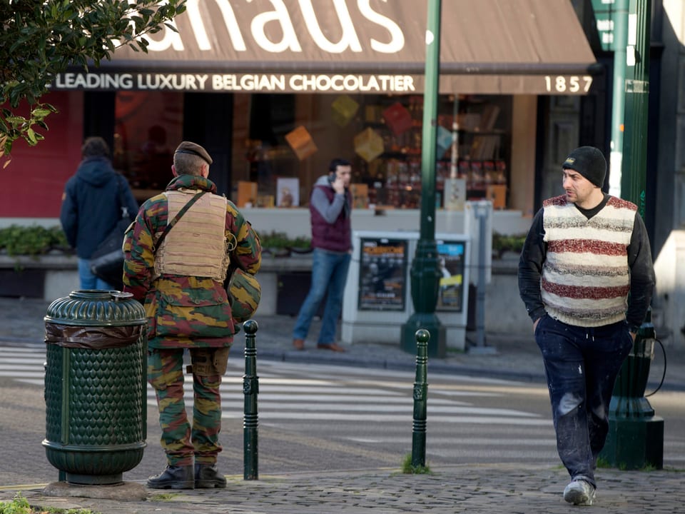 Belgischer Soldat mit dem Rücken zur Kamera beobachtet einen Schokoladeladen, ein entgegenkommender Passant mustert ihn