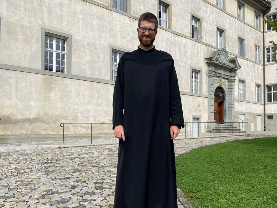Pater Philipp in langem schwarzem Gewand, lächelnd vor Klostermauer