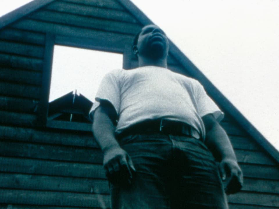 Aufnahme aus der Froschperspektive von Steve McQueen, der vor einem kleinen Holzhaus steht.