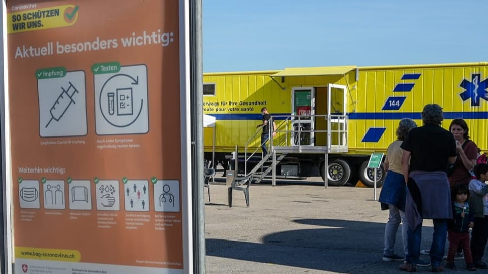 Man sieht ein Schild mit Coronaanweisungen und im Hintergrund einen riesigen grossen gelben Corona-Truck.