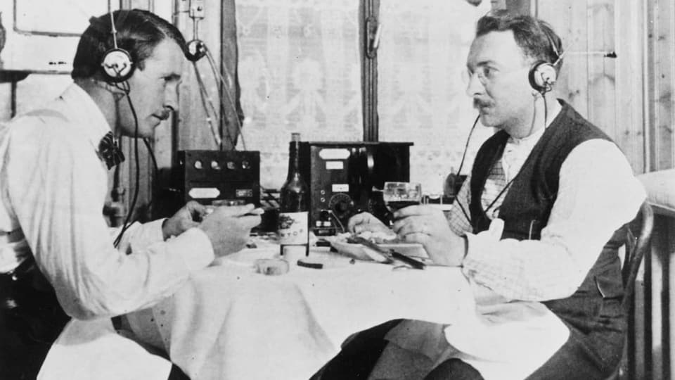 Zwei Männer sitzen um einen Radioapparat und hören mit Kopfhörern (um 1920).