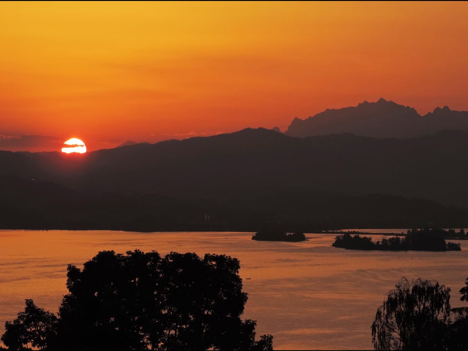 Blick auf den Zürichsee, hinter dem Hügel geht die Sonne auf. Der Himmel ist rot verfärbt.