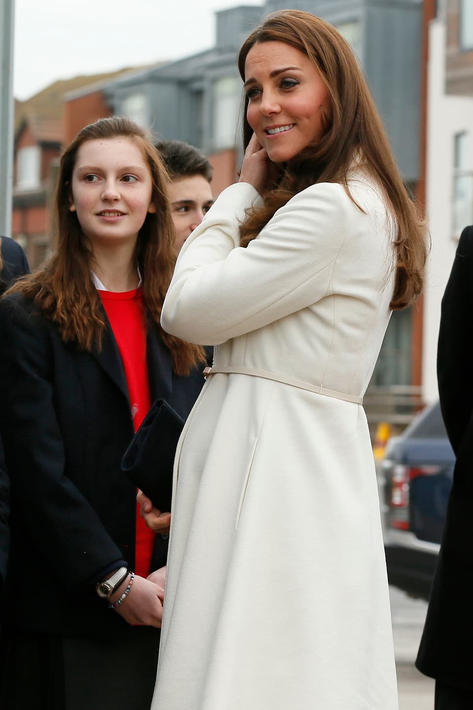 Herzogin Kate in einem weissen Mantel lächelnd und mit Mädchen sprechend.