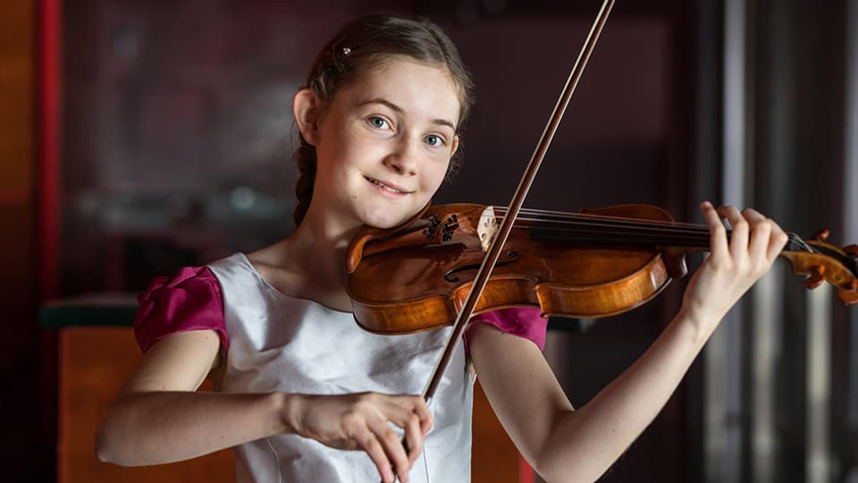 Ein Mädchen im weissen Röckchen spielt Geige und lächelt in die Kamera.