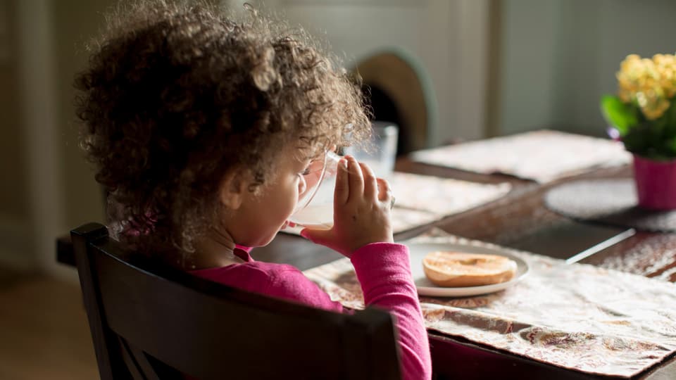 Kleines Mädchen mit lockigem Haar trinkt aus einer Tasse am Küchentisch.