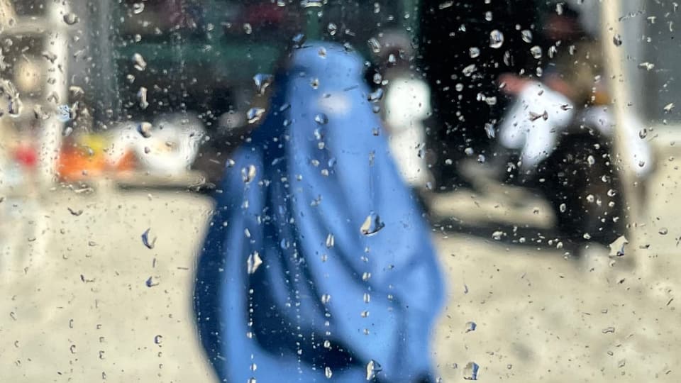 Mensch mit einer blauem Verhüllung von Kopf bis Fuss, unscharfe Figur, da hinter einem verregnetem Fenster.