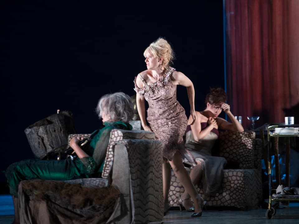 Sally Matthews (Silvia) rennt in Abendgarderobe zwischen zwei Sofas, auf denen zwei Frauen mit zu Boden gerichtetem Blick sitzen.