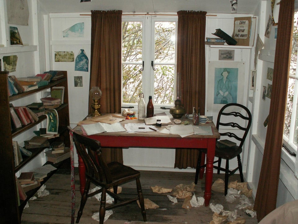 Ein Zimmer mit Arbeitstisch, Stühlen und einem Bücherregal.