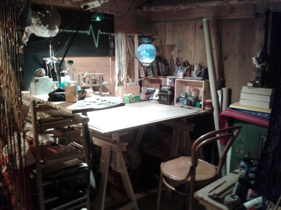 Schreibtisch mit ganz vielen Sachen drauf in einer Art Holzkammer.