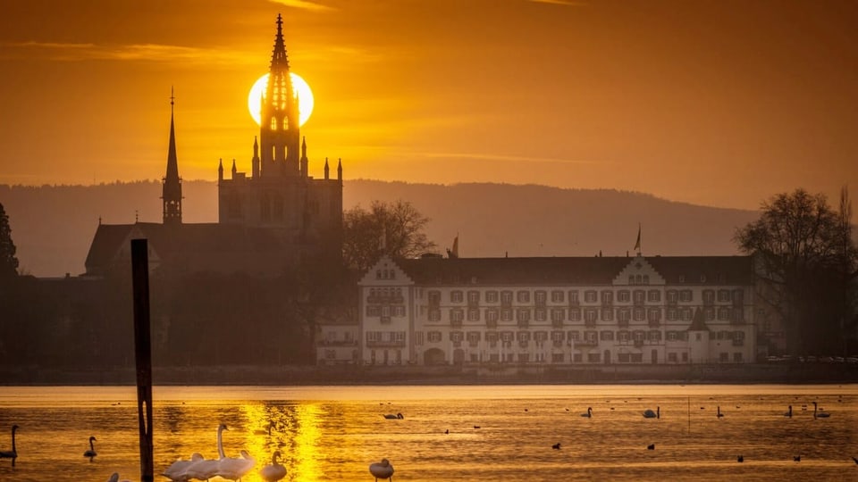 Eine Kirche und ein altes, langes Gebäude am See mit Schwänen bei orangem Sonnenuntergang.