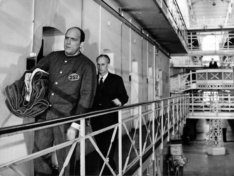 Eine Szene im Gefängnis. Ein Mann in Häftlingskleidung läuft den Gefängniszellen entlang. Hinter ihm folgt ein Mann im Anzug.