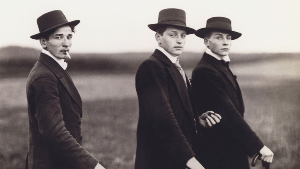 Drei Männer auf einem Foto. Sie tragen alle Hut und halten einen Stock.