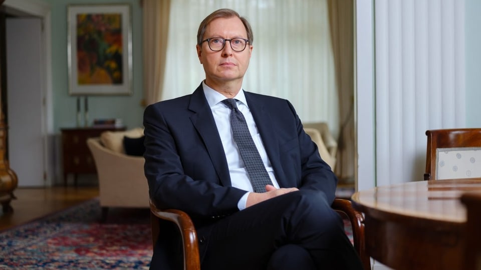 Michael Flügger sitzt auf einem Stuhl. Gesichtsausdruck neutral, Krawatte und Anzug. Ganz diplomatisch, auch bebrillt.