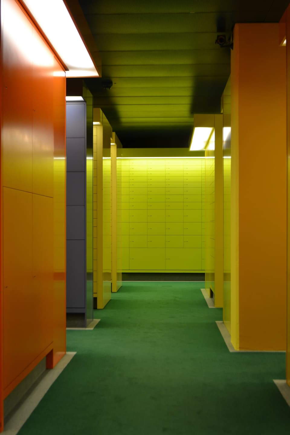 Ein Gang mit grünem Teppich, daneben orangefarbene Wände, am Ende des Gangessind Tresorfächer sichtbar.