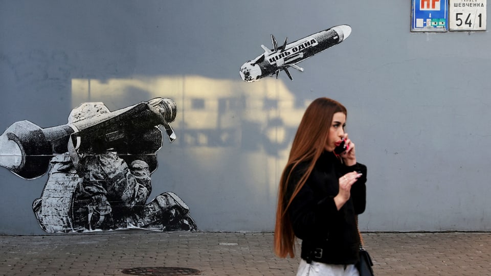 Eine junge Frau steht telefonierend vor einem Grafitti an einer Hauswand, das einen Soldaten zeigt