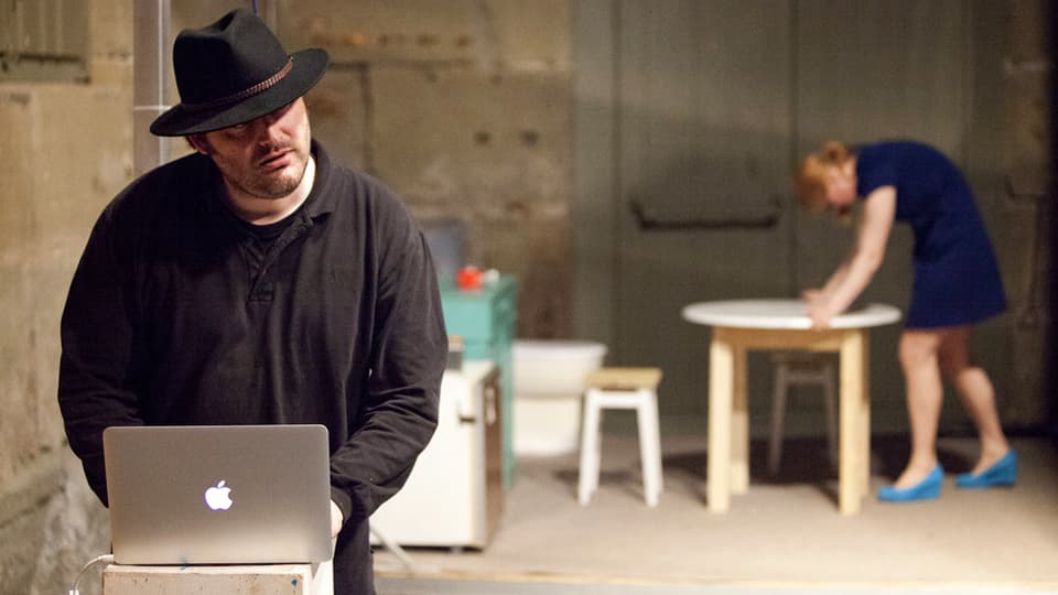 Ein Mann mit Hut und dunklem Pullover steht an einem aufgeklappten Laptop.