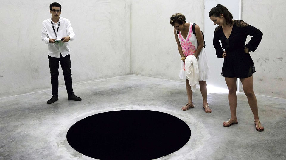 Im Museum: Drei Leute schauen auf ein schwarzes Loch im Boden.