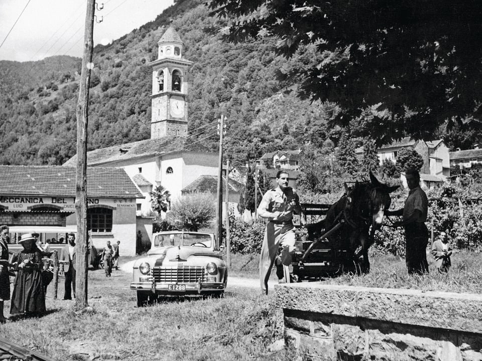 Filmszene in Schwarzweiss: Tessiner Dorf mit einer steinernen Kirche. Ein Mann rennt neben einem Pferdewagen. Im Hintergund ein altmodisches Auto mit offenem Verdeck. 