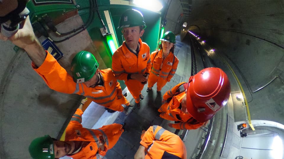 Männer und Frau, in orangen Arbeiter-Schutzanzügen und mit Helm, Fish-Eye-mässig verzerrt weil von oben fotografiert.