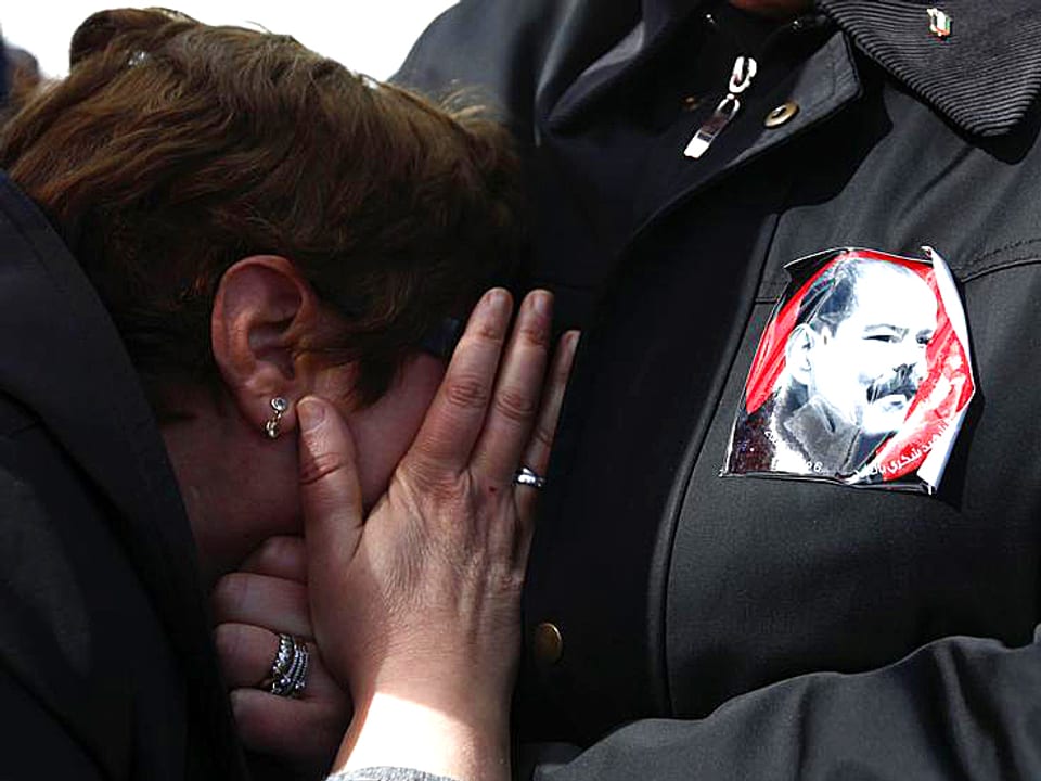 Eine Frau weint heftig an der Brust ihres Mannes. Dieser trägt ein Abziehbild des verstorbenen Oppositionspolitikers an seinem Revers.
