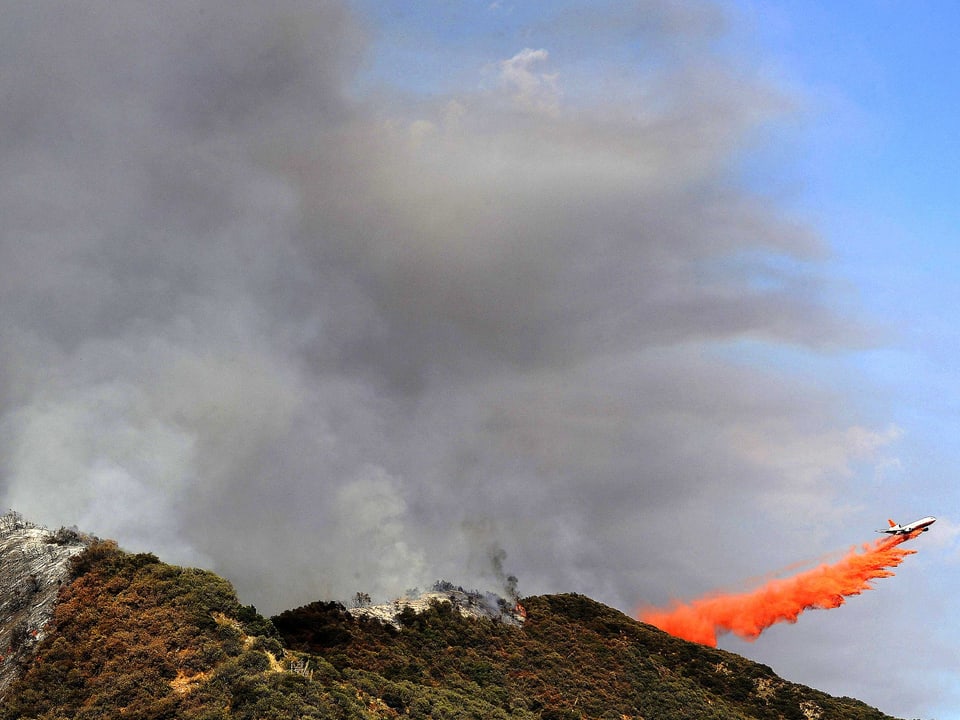 Ein Löschflugzeug lässt orange Löschflüssigkeit über einen brennenden Bergkamm fallen.