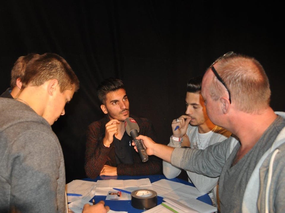 Junge Männer stehen um einen Tisch, der Reporter hält einem das Mikrofon vor den Mund.